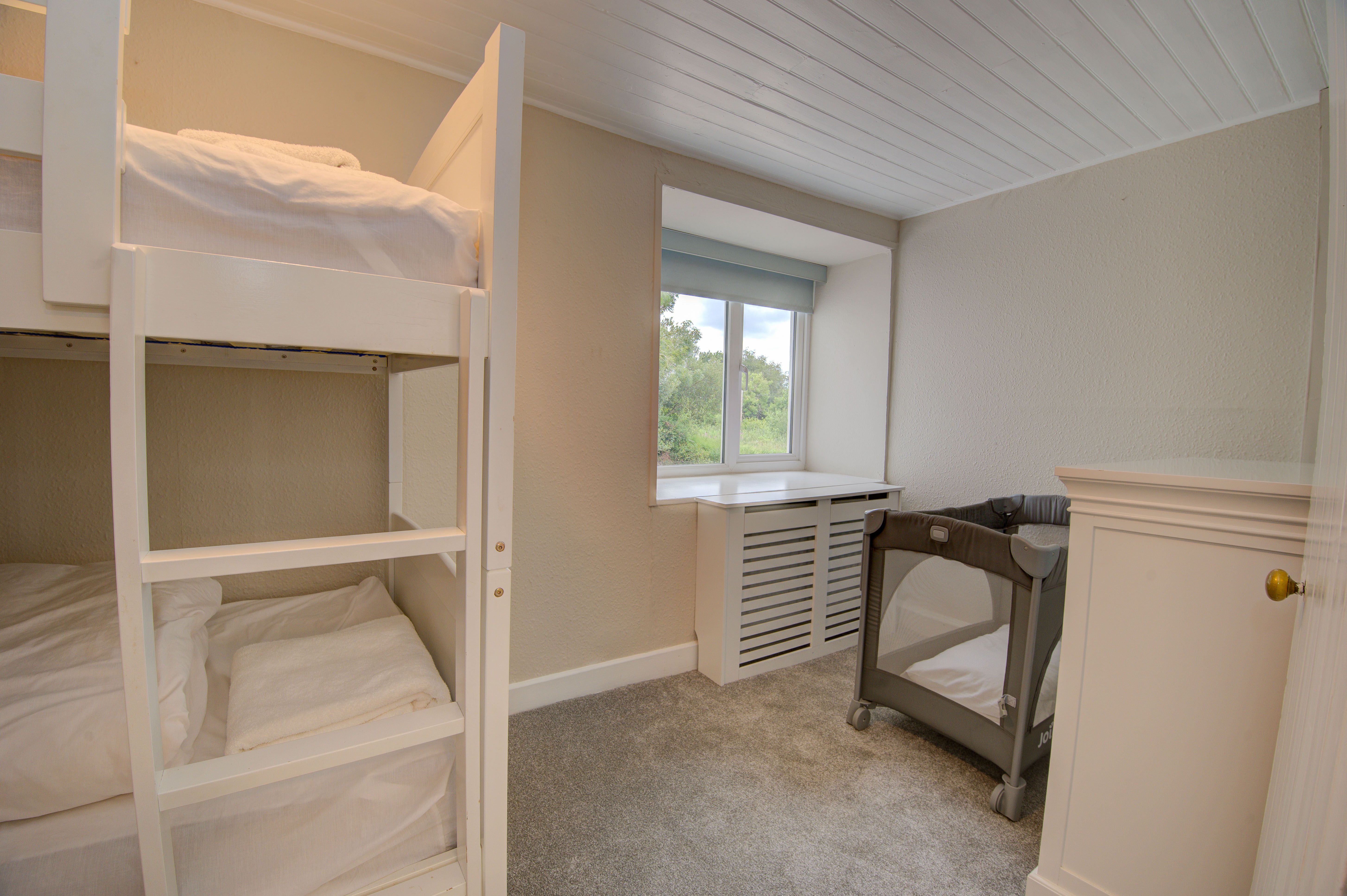 The cottage bunk room, with a white bunk bed and a grey travel cot by the window
