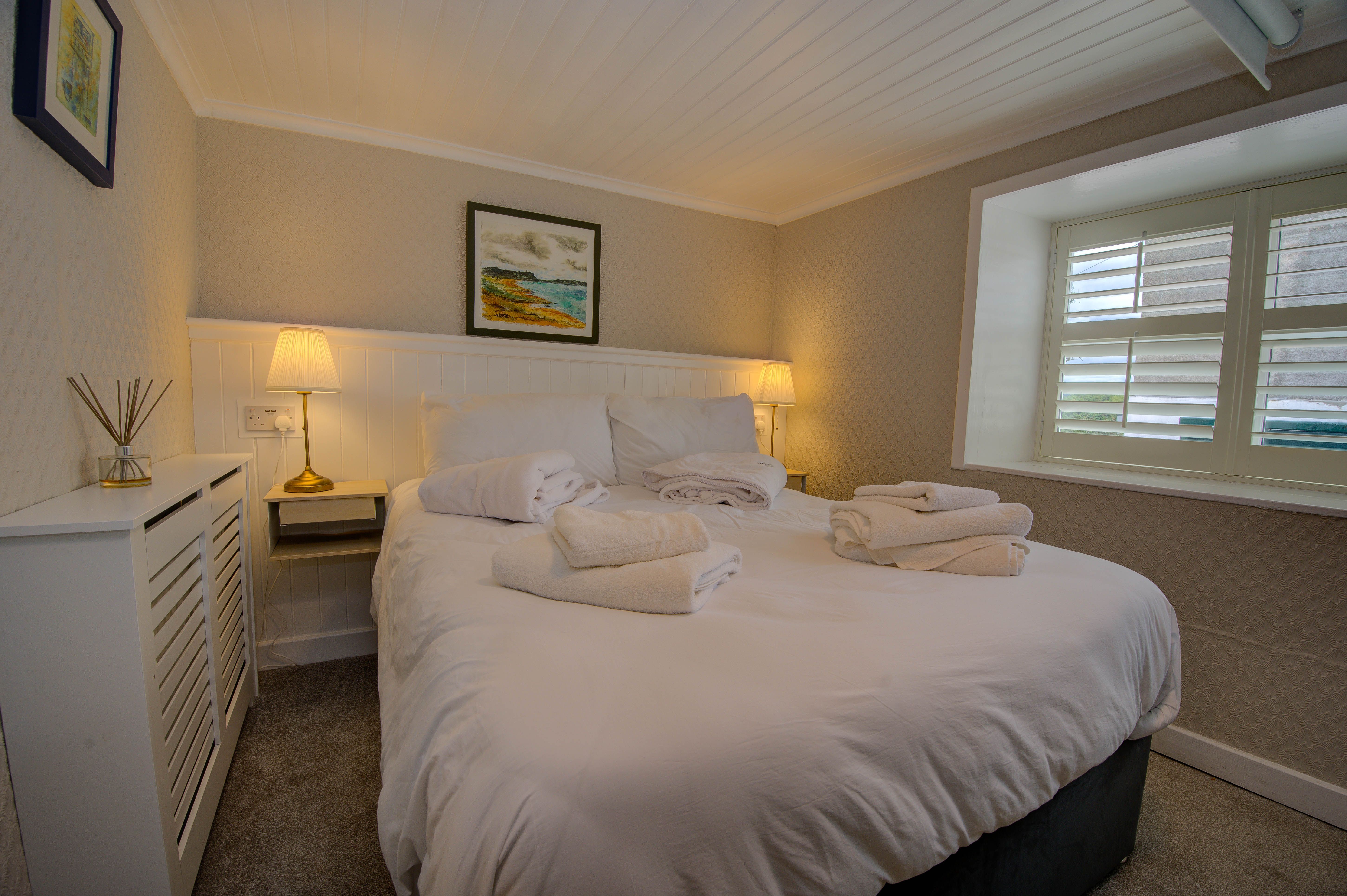 The cottage main bedroom lamp-lit, shuttered window letting in low light