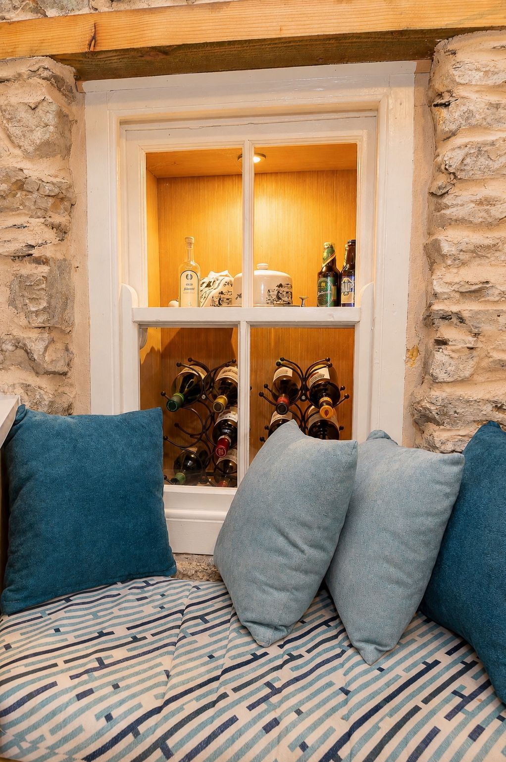 The finished window seat and wine store — the little window boxed out and lit, bottles racked behind, a cushioned seat built around it in the stone wall.