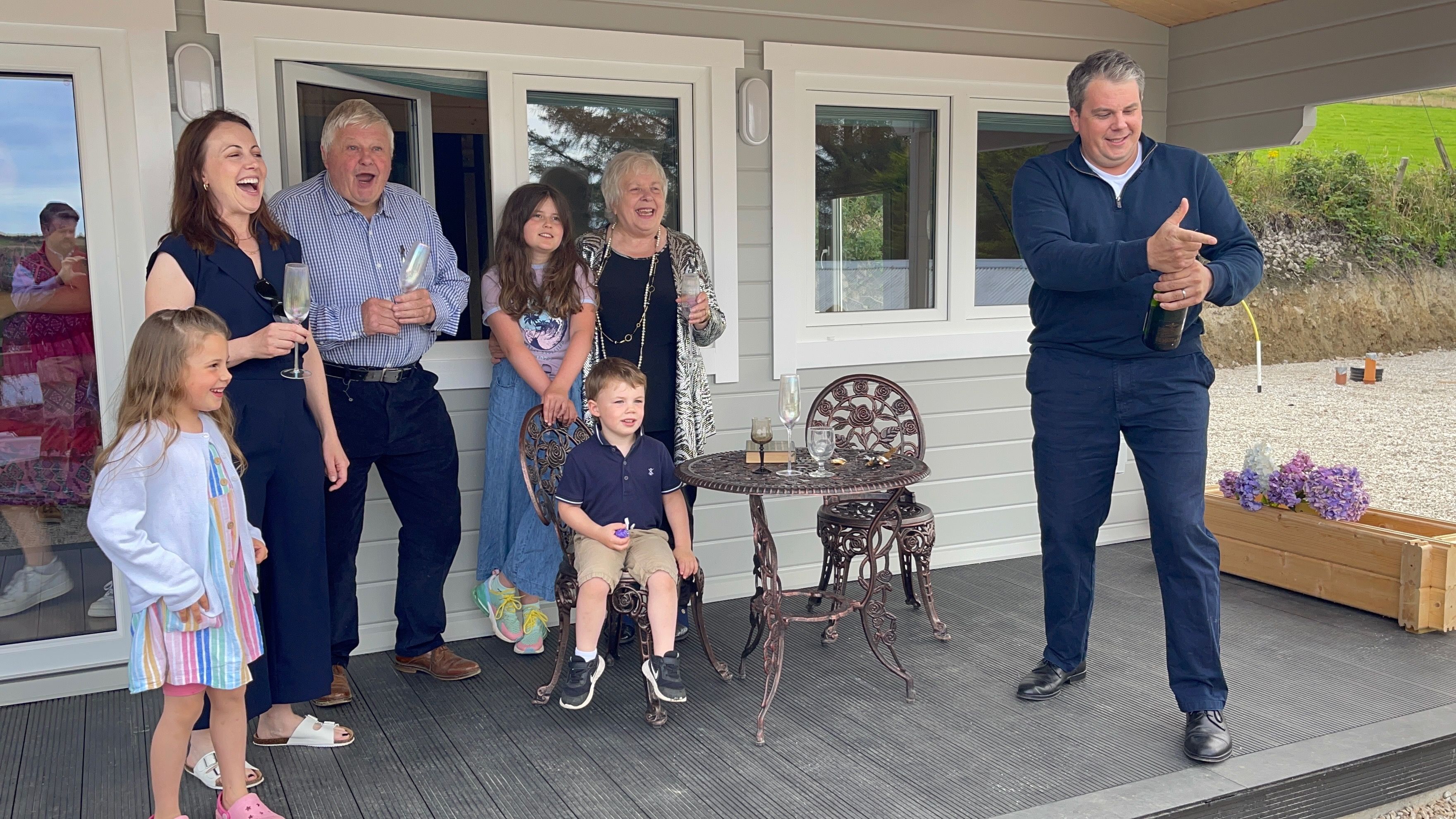 The family gathered on the sunlit deck at Rae's Cabin, mid-toast as a bottle of sparkling wine is opened, with three young children among them.