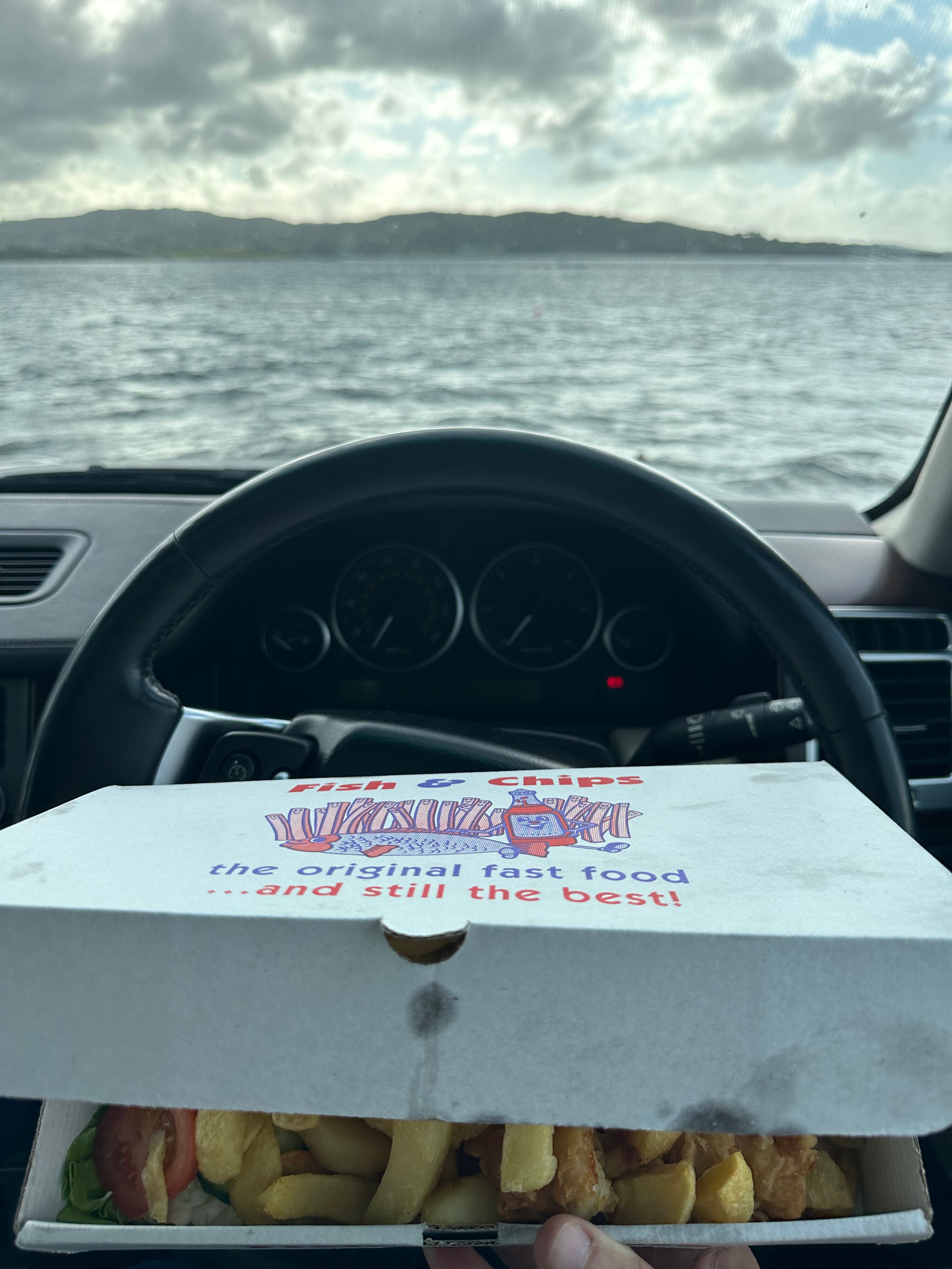 An open box of golden fish and chips on a car dashboard, with the calm water of Mulroy Bay and the far shore through the windscreen beyond