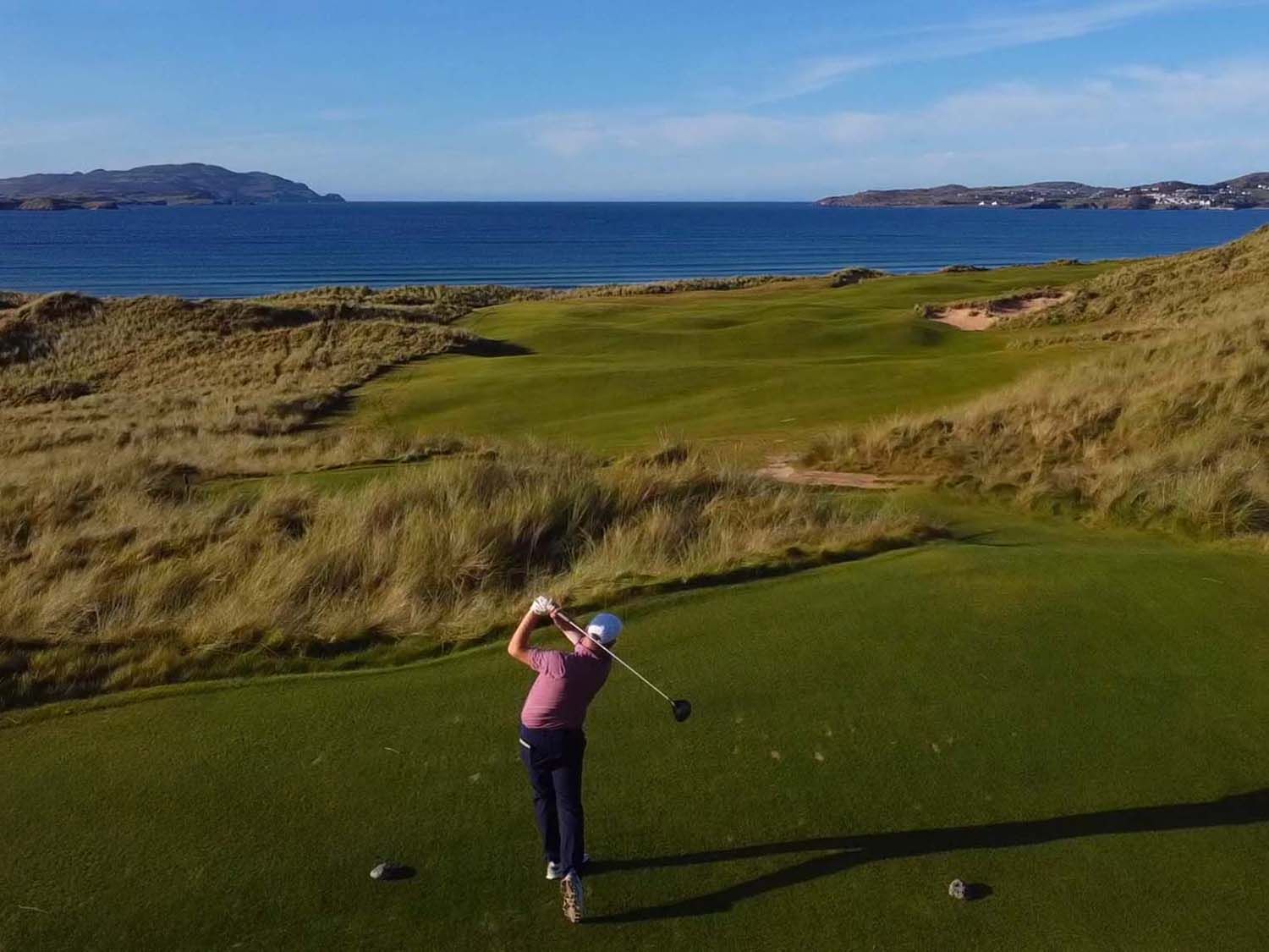 A golfer at the top of the follow-through on an elevated tee at St Patrick's Links, long shadow in low evening sun