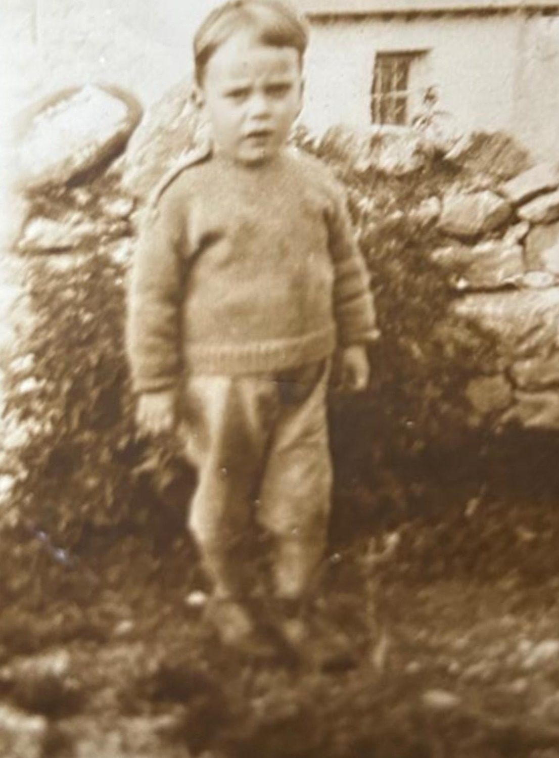 An aged sepia photograph of Davey John as a young boy, in a hand-knitted woollen jumper and short trousers, standing by a dry-stone wall with the whitewashed cottage behind him.