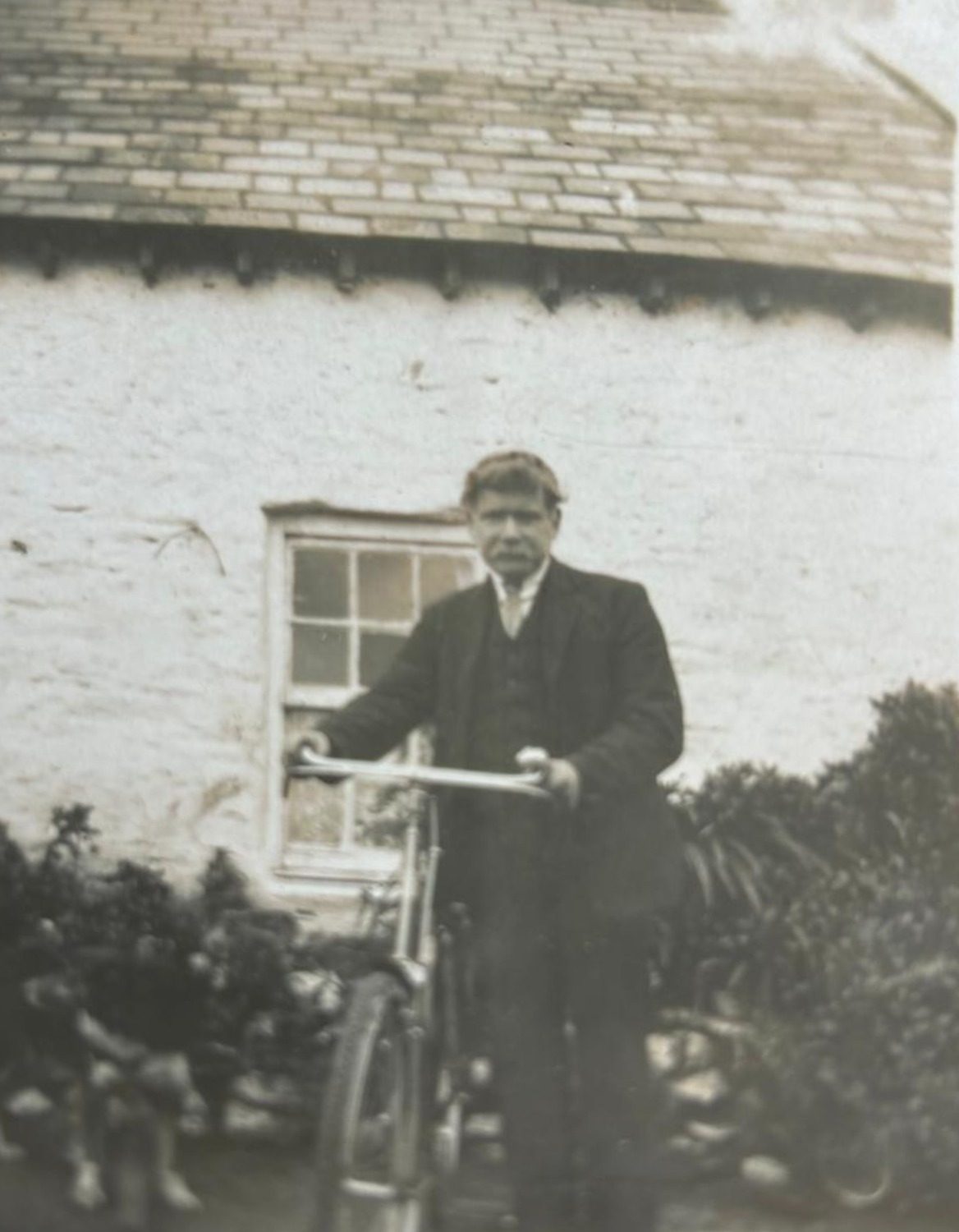 An aged black-and-white photograph of James Graham, in a dark three-piece suit and flat cap, standing astride a bicycle in front of the whitewashed cottage at Devlinmore.