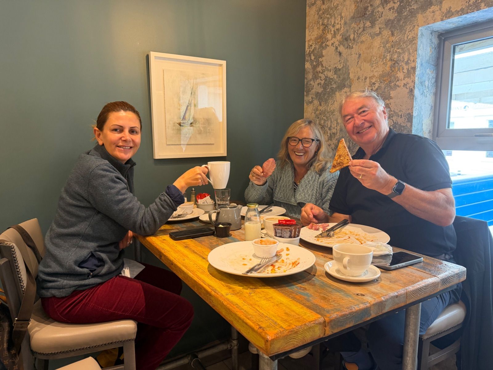 Three people smiling at a wooden café table with mostly cleared breakfast plates, cups, and a small teapot in front of them. A framed watercolour print of a sailing boat hangs on the teal wall behind them, and a bright window is visible to the right.