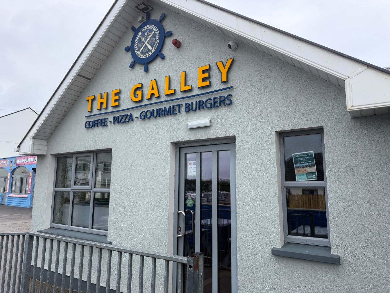 The exterior of The Galley café in Downings, a white rendered building with large yellow and blue lettering reading 'THE GALLEY' and a ship's wheel logo, advertising coffee, pizza and gourmet burgers. A colourful neighbouring shopfront is visible to the left.