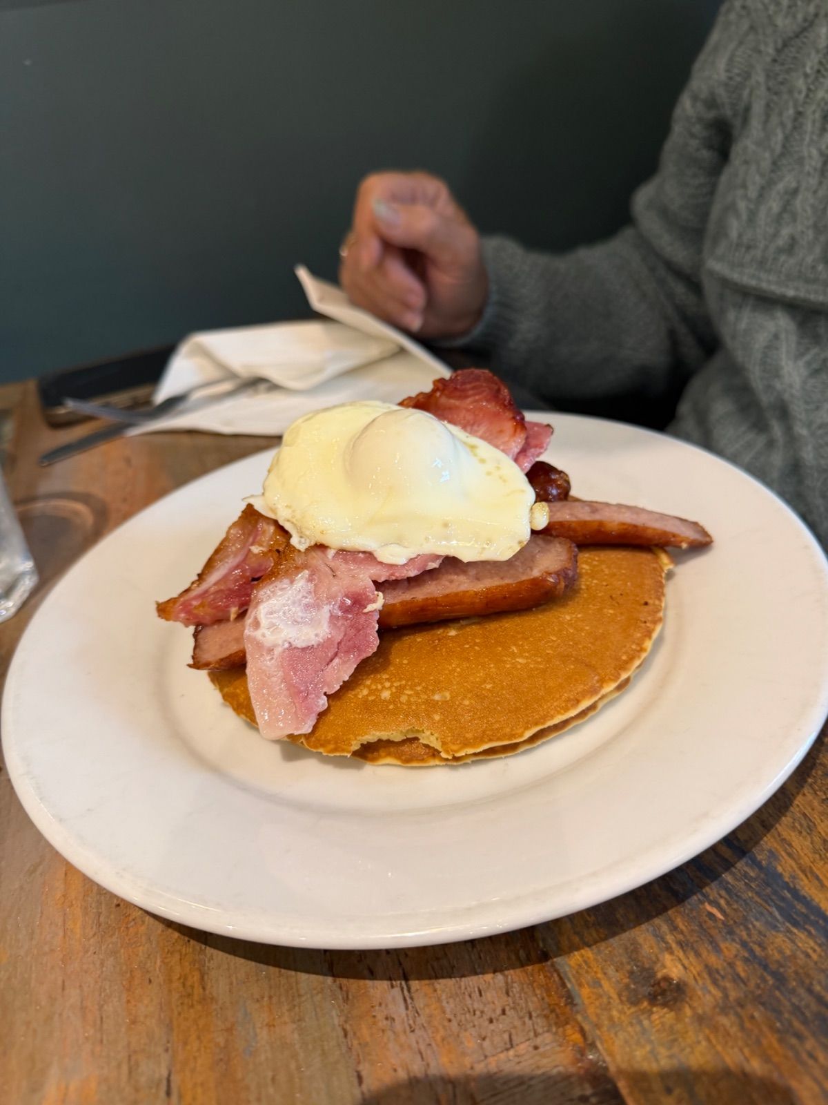 A white plate on a wooden café table holding a golden pancake topped with rashers of back bacon, lengthways-sliced sausage and a single egg with a soft yolk. A person in a grey cable-knit jumper is visible out of focus in the background.