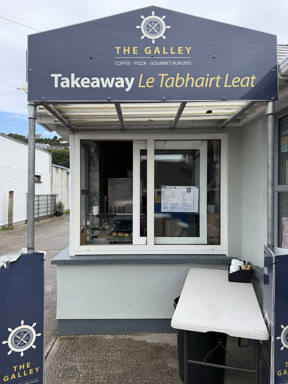 A takeaway hatch set into a pale grey-green wall beneath a navy canopy reading 'The Galley — Coffee, Pizza, Gourmet Burgers' and 'Takeaway, Le Tabhairt Leat', with a ship's wheel emblem above. A glass case of pastries stands on the counter inside, and a narrow white ledge with napkins and cutlery sits to the right.