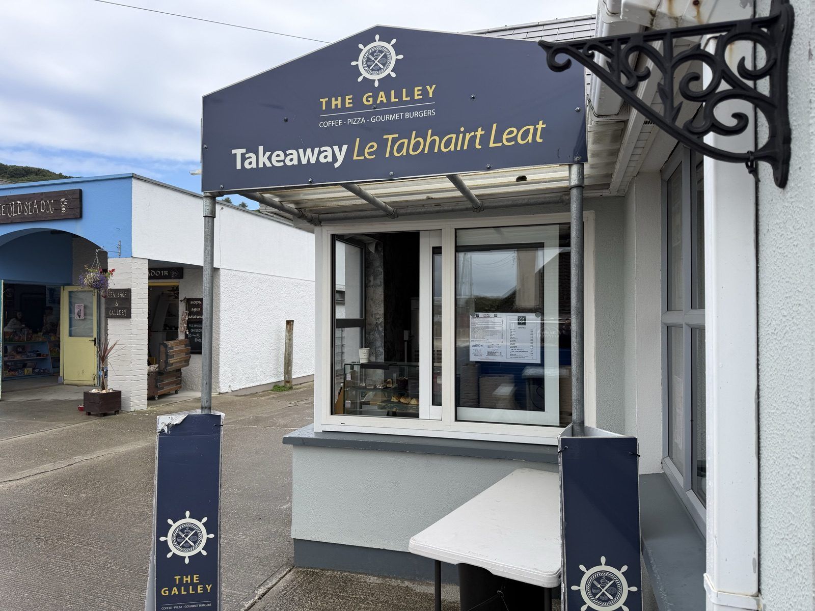 The same takeaway hatch seen from along the street on an overcast day, its navy canopy and ship's wheel sign above a sliding window, with two matching navy pavement signs in front. The blue frontage of the Old Sea Dog stands next door to the left, a wooden sign above its archway.