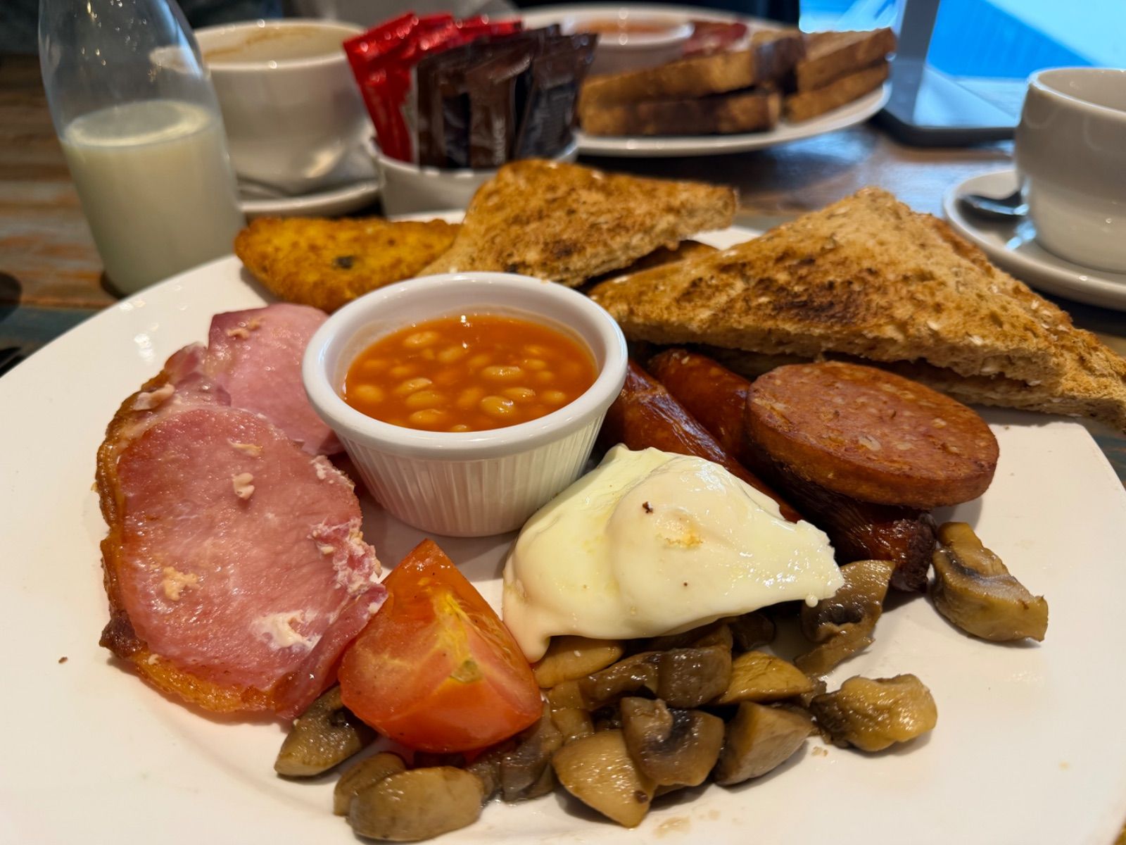 A full Irish breakfast plate with bacon, a poached egg, sausage slices, mushrooms, grilled tomato, baked beans in a ramekin, a potato cake, and brown and white toast triangles on a wooden café table.