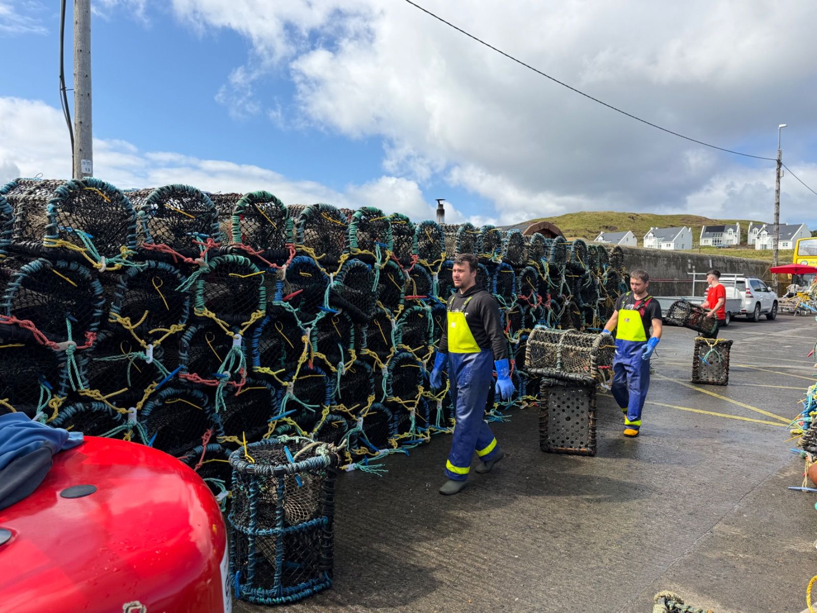 Two fishermen in yellow-and-blue waterproof bibs and blue rubber gloves carry lobster pots across a harbour pier alongside a tall stack of pots. A third worker in a red top is visible further back. Houses on a green hillside sit behind a harbour wall in the background under a partly cloudy sky.