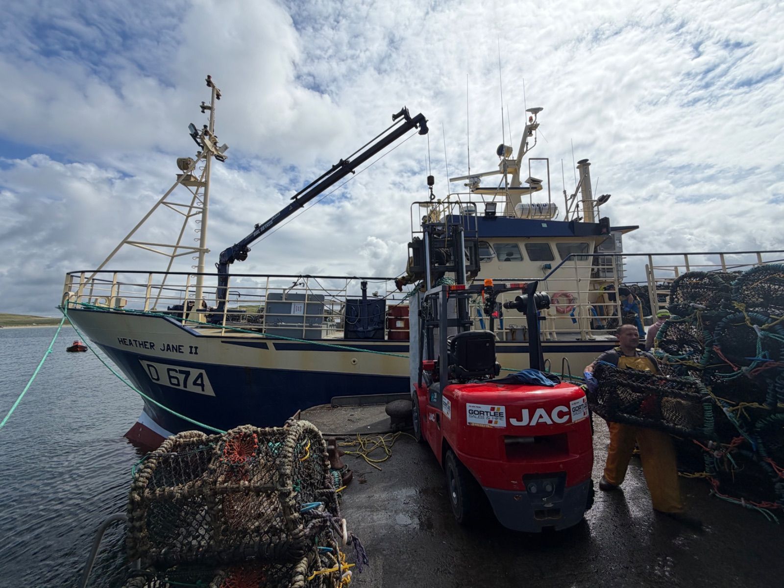 The full side profile of the Heather Jane II, D 674, moored at a wet harbour pier. A red JAC forklift is parked close to the hull and a fisherman in yellow waterproofs handles a lobster pot in the foreground. Stacks of pots sit on the quayside in the foreground and background under a bright cloudy sky.