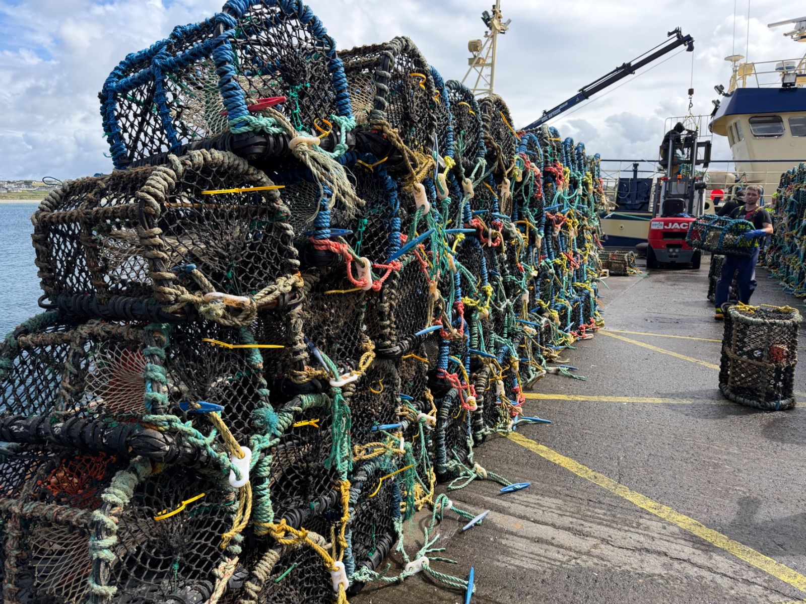 A long, close-up wall of stacked crab pots on a harbour pier, their mesh frames woven through with coloured ropes in teal, yellow, red, and blue. In the background, the superstructure of the Heather Jane II and a red forklift are visible, with a crew member carrying a pot.