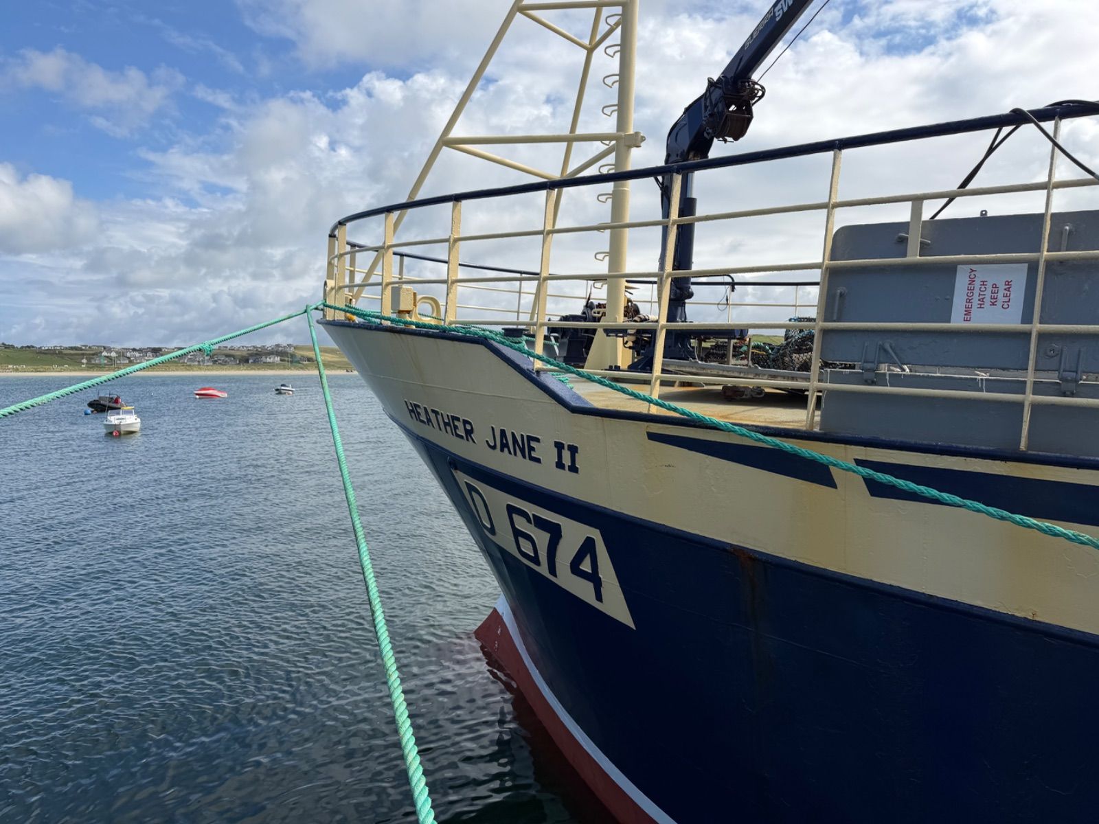 The navy-and-cream bow of the fishing vessel Heather Jane II, registration D 674, moored at a pier with green rope lines, smaller boats visible on calm water behind it and a village on the far shore.