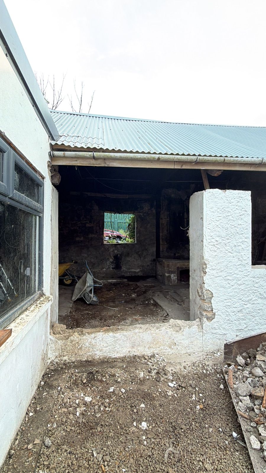 Exterior view of the forge building mid-clearance, showing the open front with no door, a corrugated metal roof, a whitewashed rendered pier to the right of the opening, two wheelbarrows inside, rubble and dug earth in the foreground, and a whitewashed building to the left.