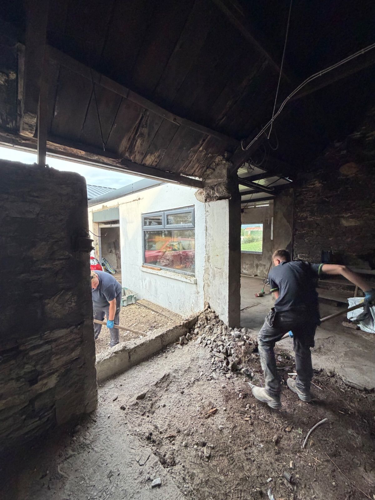 Two workers inside the forge shovelling rubble and debris near a broken-out doorway opening, with exposed dark timber ceiling beams above, a rough stone pillar in the foreground, a whitewashed building and window visible outside, and a pile of broken masonry on the ground.
