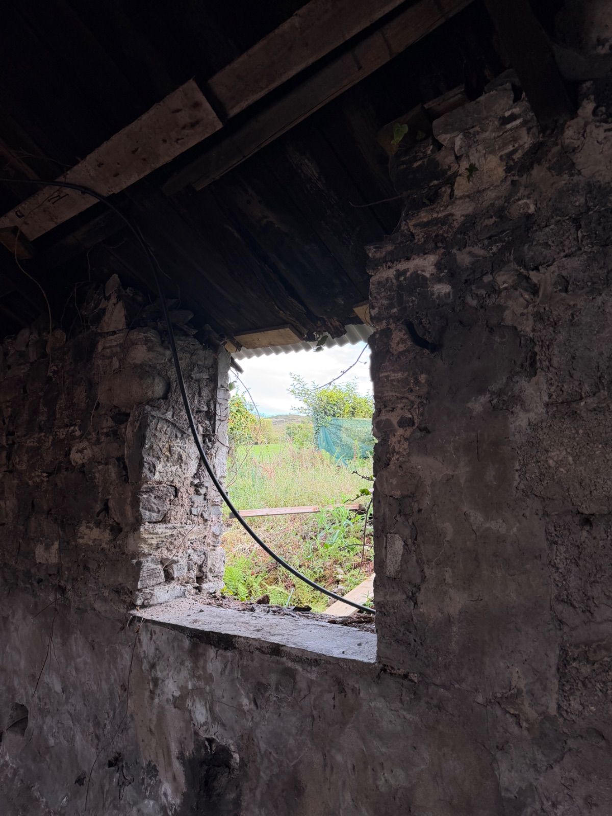 View from inside the dark interior of the forge looking out through an unglazed stone window opening, with green fields, hedgerows, and overcast sky visible beyond, a black pipe running across the sill, and rough stonework forming the reveal on all sides.