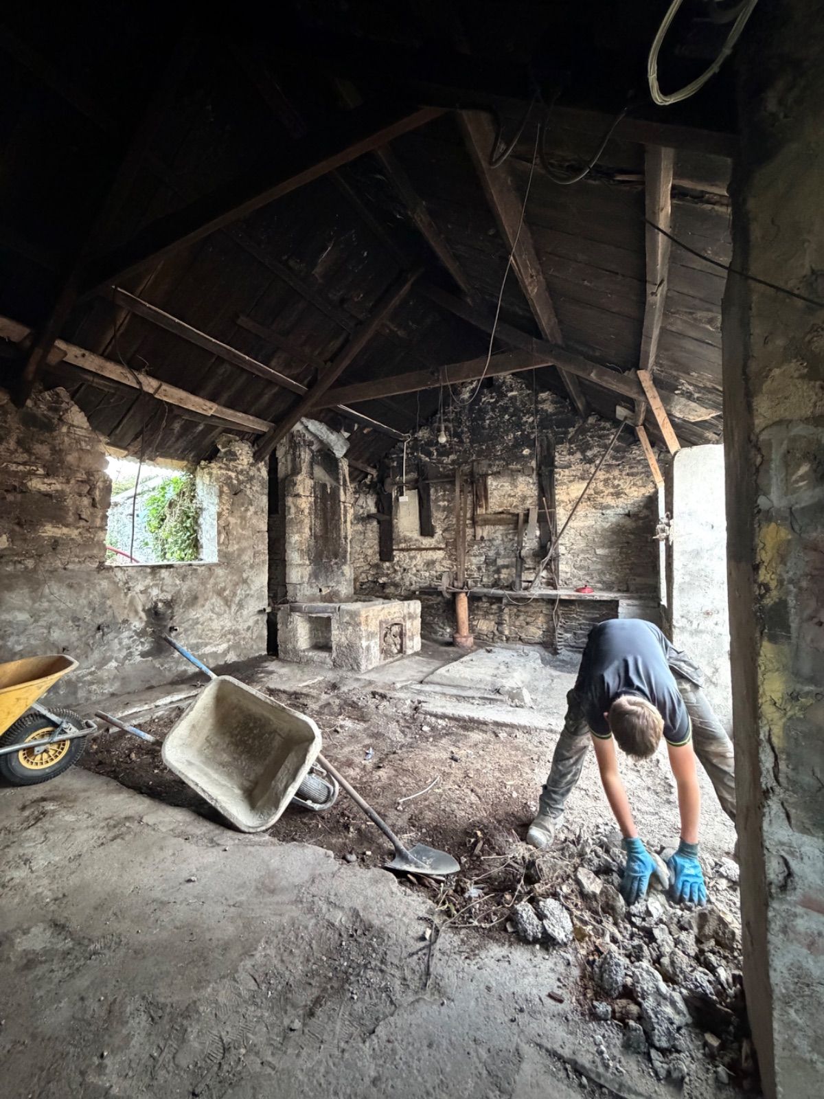 Interior of the forge with a person bent over picking up rubble near two wheelbarrows, the stone hearth and chimney arch visible in the background, rusted ironwork on the back wall, exposed timber roof trusses above, and a small window opening letting in light from the left.