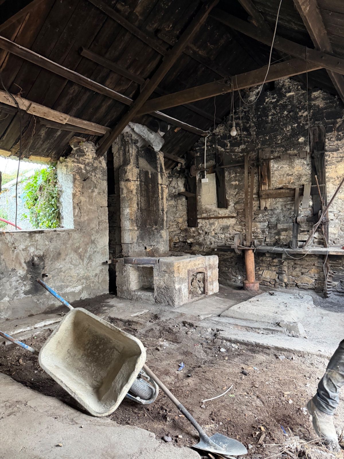 Interior of the forge looking toward the stone hearth and chimney arch, with rusted iron machinery and a wooden workbench along the back wall, exposed timber roof trusses above, a bare bulb hanging on a wire, and a wheelbarrow and shovel in the foreground.