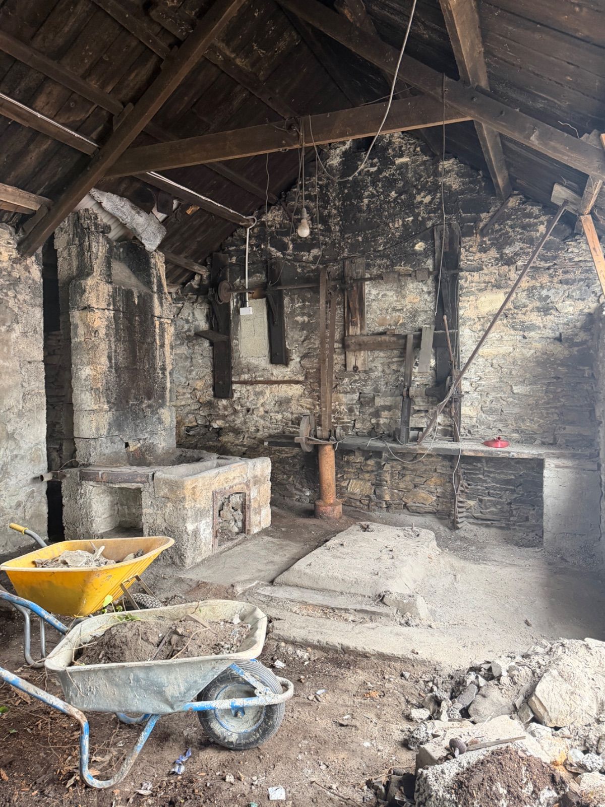 Interior of the old blacksmith's forge during clearance work, showing the stone hearth and chimney arch, rusted ironwork machinery on the back wall, exposed timber roof trusses blackened with age, two wheelbarrows in the foreground, and rubble on the earthen floor.