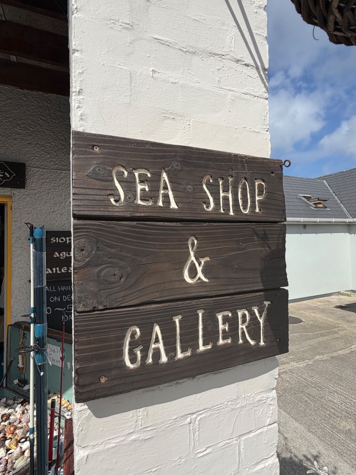 A close-up of a dark wooden sign reading 'Sea Shop & Gallery' in carved white lettering, mounted on a white-rendered corner pillar in bright sunshine.