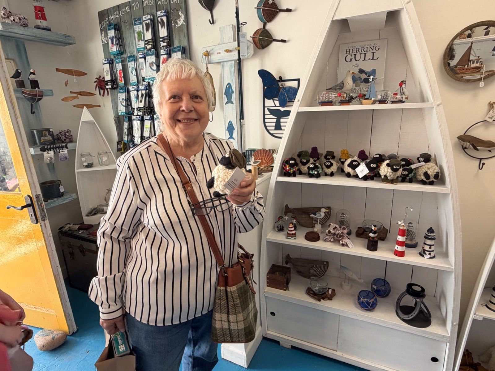 A smiling older woman with short white hair stands inside the Old Sea Dog shop, holding a small felted sheep ornament and looking at the camera. Behind her is a white boat-shaped display unit filled with felted sheep, lighthouse ornaments, and other maritime gifts.