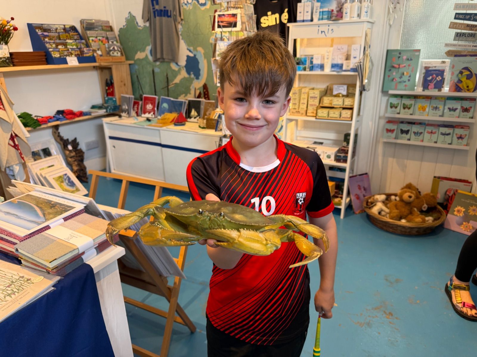 A young boy in a red and black number 10 football shirt grins at the camera while holding up a large realistic-looking fake green crab inside the Old Sea Dog shop, with bookshelves and gift displays visible behind him.