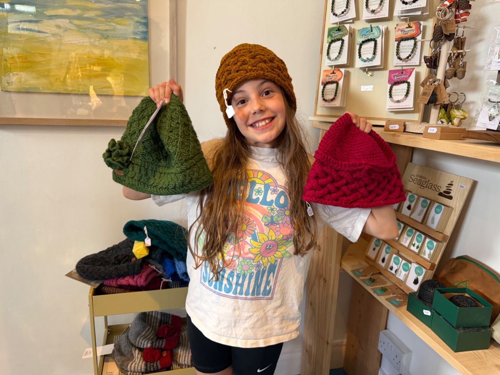 A young girl wearing a rust-coloured cable-knit hat smiles broadly at the camera while holding up a dark green knitted hat in her left hand and a red one in her right, inside a shop with more hats stacked on a rack behind her and gemstone jewellery cards on shelving to the right.