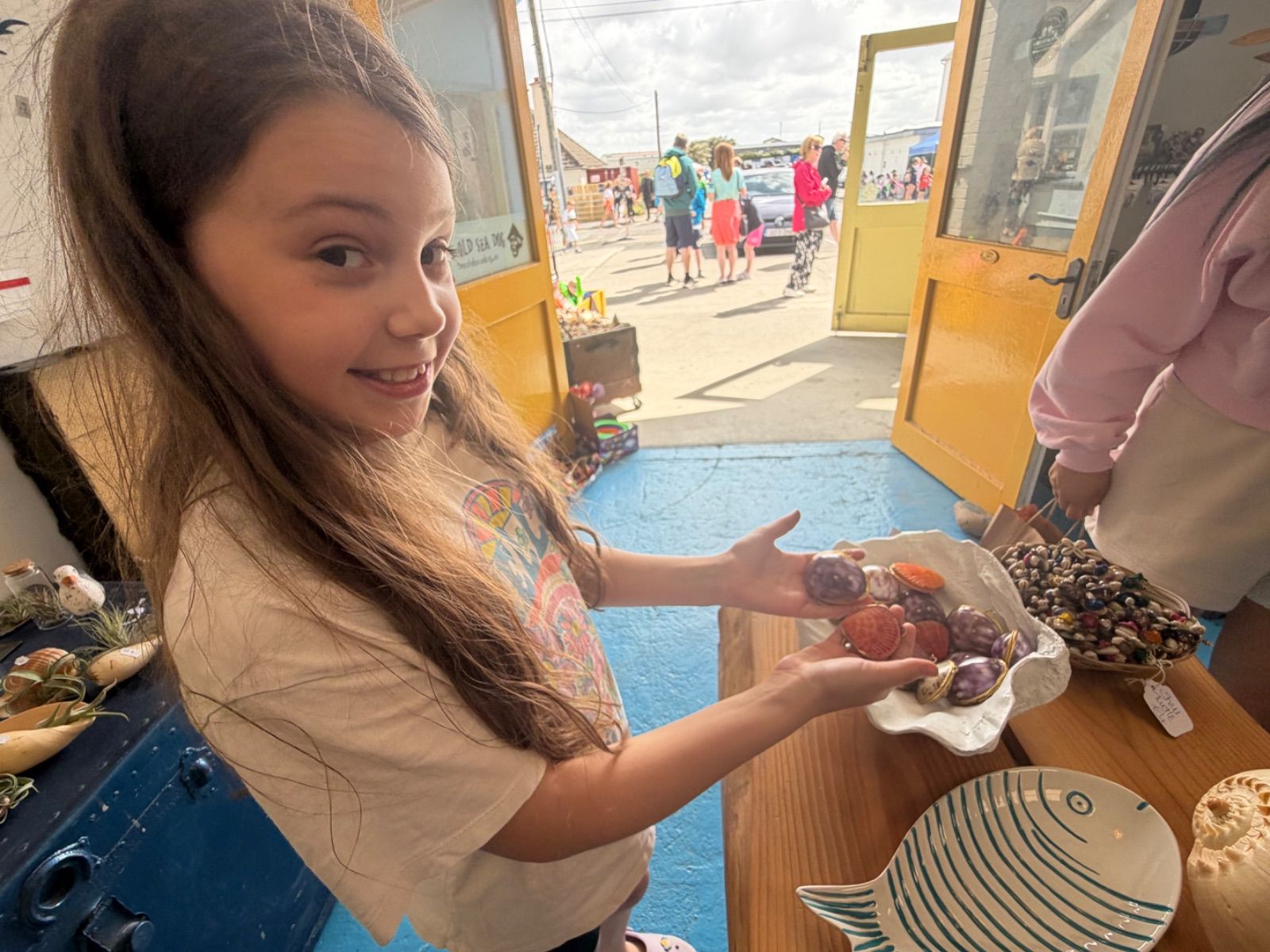 A young girl with long brown hair glances back at the camera with a slight smile while holding out a ceramic shell-shaped dish containing polished shells in orange, red and purple tones, several of them mounted with small gold clasps as pendants; the yellow doors of the Old Sea Dog are open behind her showing a busy street outside.
