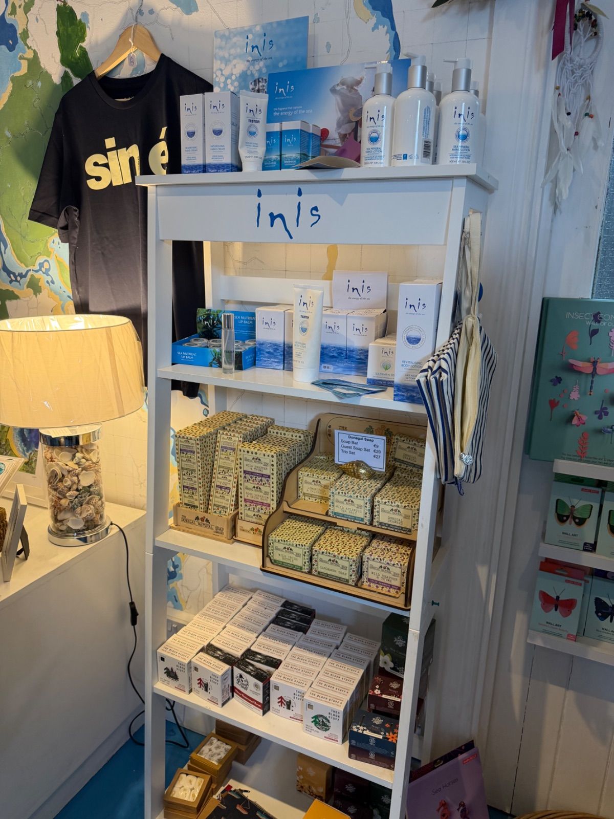 A tall white shelving unit inside the Old Sea Dog displaying multiple Inis branded fragrance and skincare products on the upper shelves, Donegal Soap sets in the middle, and The Black Pepper Company soap boxes on the lower shelves; a black t-shirt with 'Siné' printed on it hangs to the left, and a map covers the wall behind.
