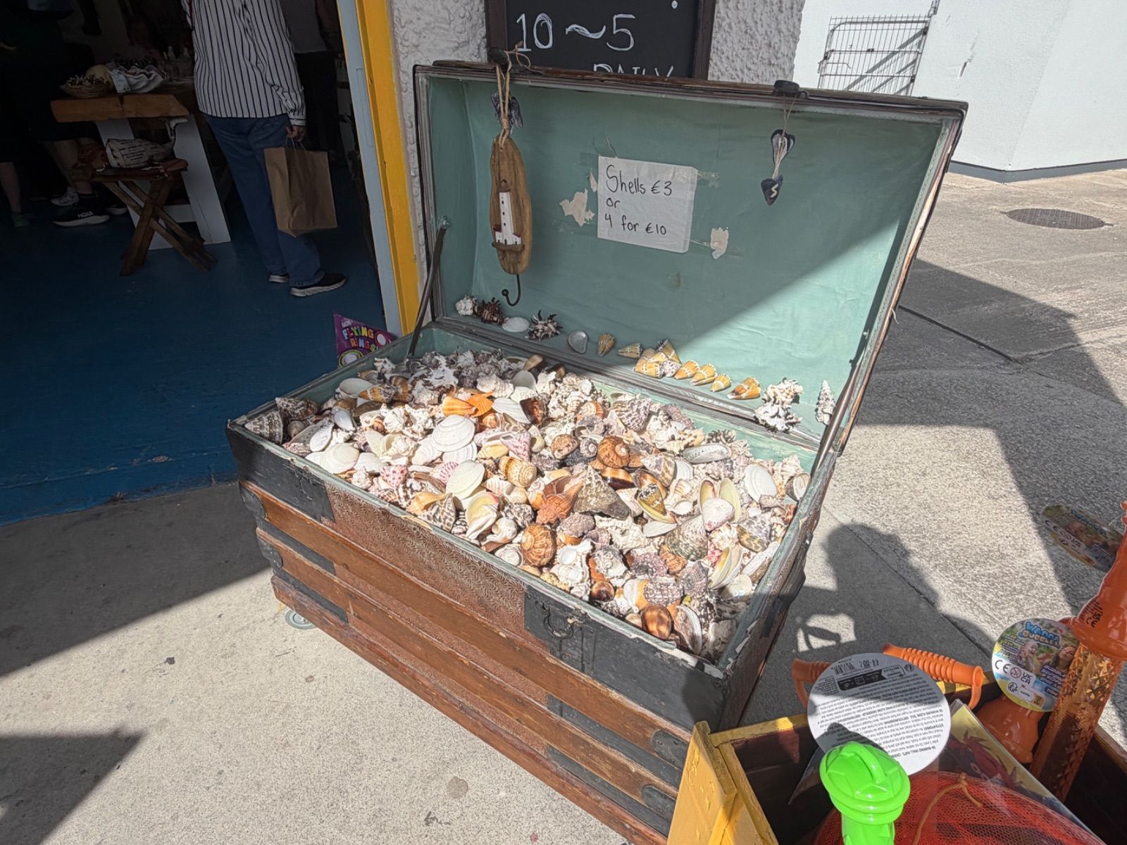 An old wooden trunk with metal banding sits open on a sunlit pavement outside the Old Sea Dog, filled with a large mixed collection of shells of various sizes, shapes and colours, with a handwritten sign inside reading 'Shells €3 or 4 for €10'.
