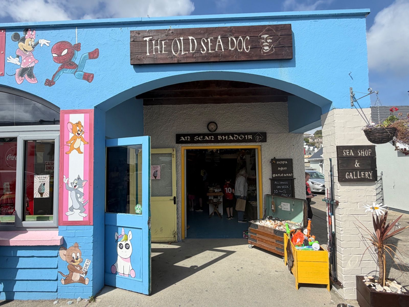 The bright blue exterior of the Old Sea Dog shop in Downings, with cartoon characters painted on the walls, a dark wooden sign above the arched entrance, and a yellow door open wide to the street.