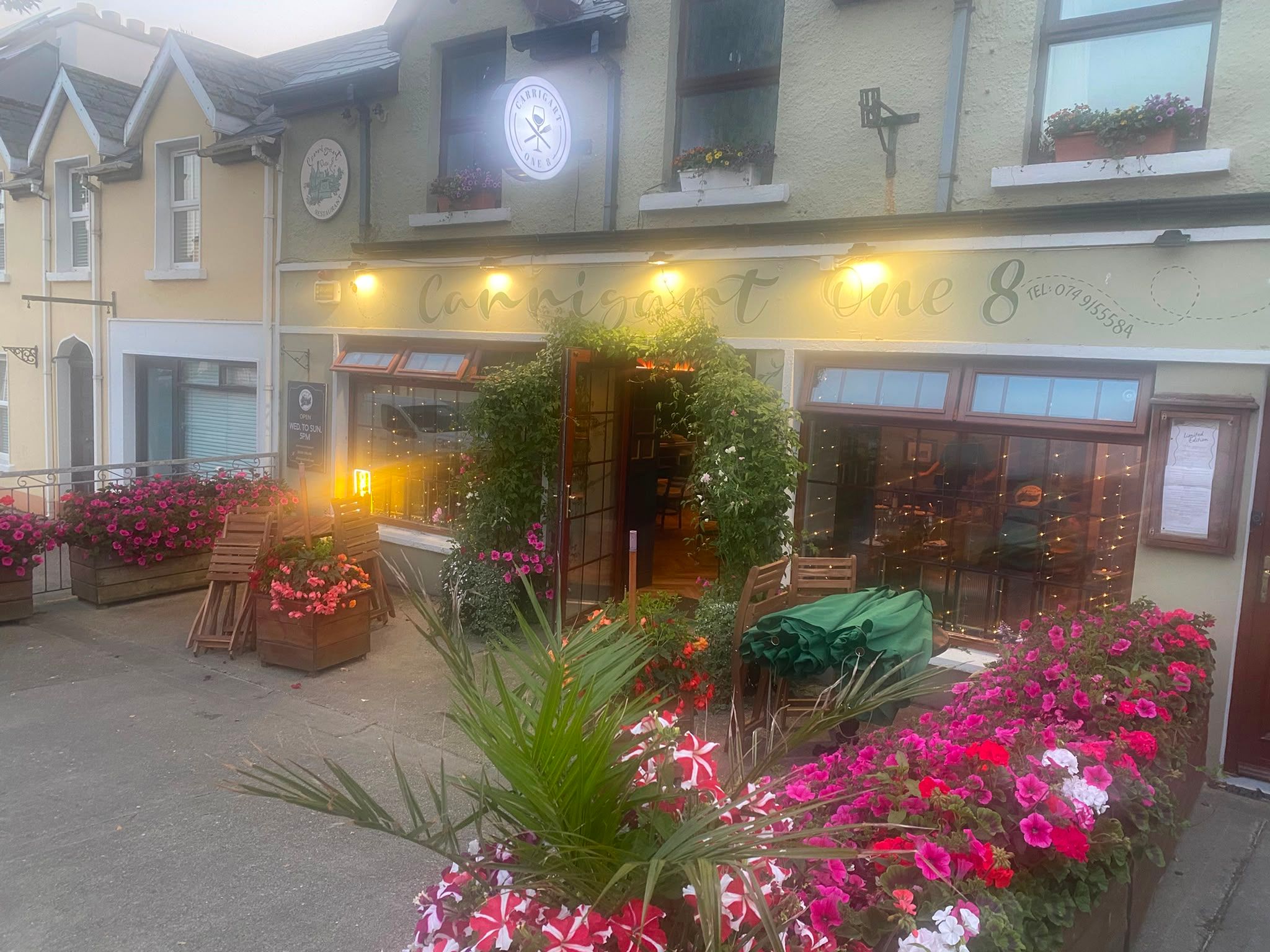 The Carrigart One 8 restaurant on Main Street at dusk: a cream terraced frontage with hand-painted lettering across the fascia, warm lights above the windows, an ivy arch around the open front door and planters of petunias along the terrace rail.