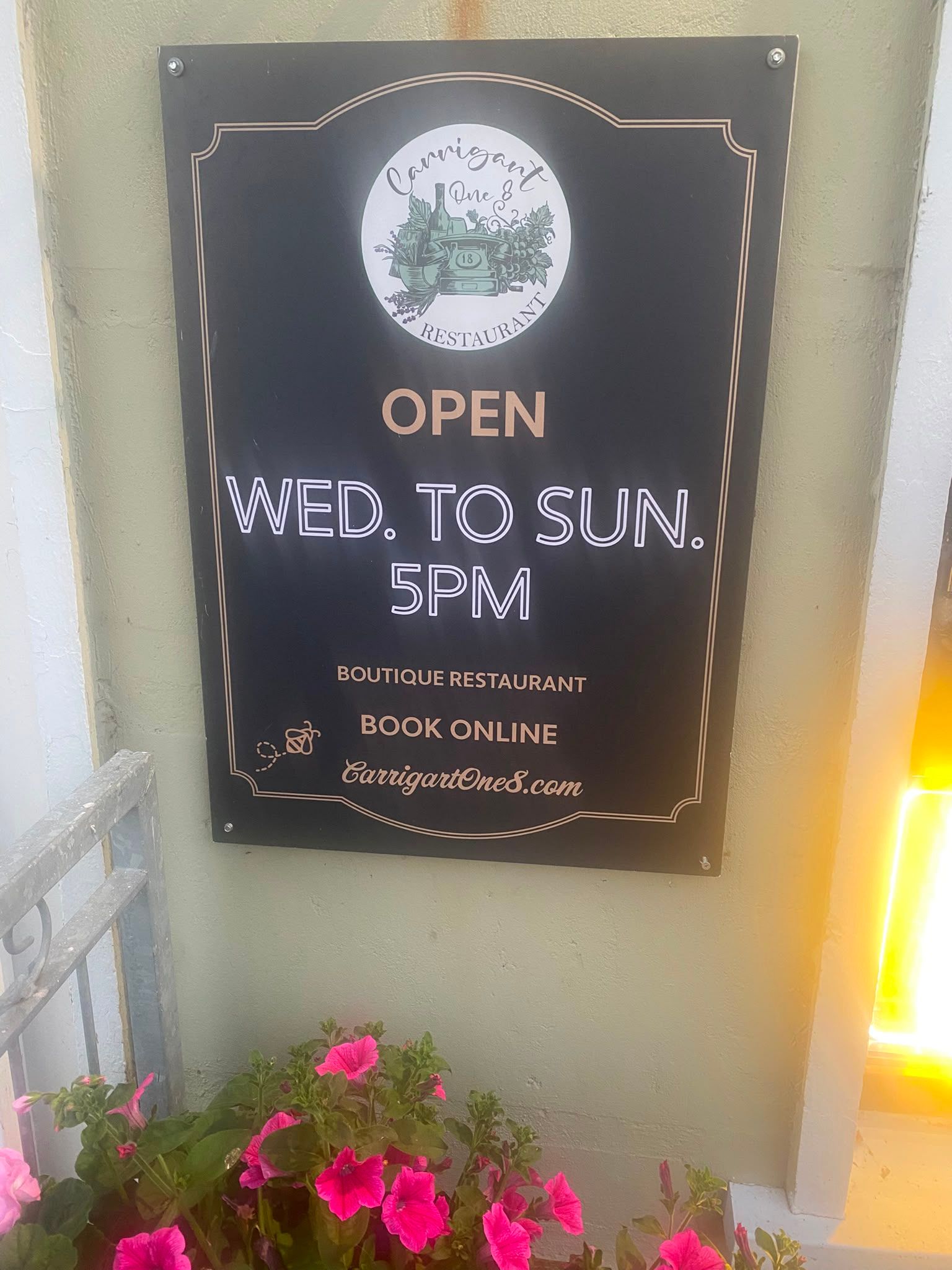 The Carrigart One 8 wall sign in evening light: a black board with the round restaurant crest, open Wednesday to Sunday from 5pm, on a sage-green wall above a window box of pink petunias.