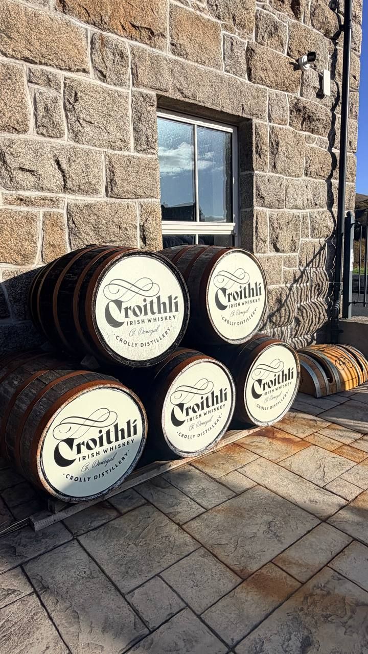Whiskey barrels stacked on a pallet against the granite wall of the old barracks, in low sun, with a window above and stamped paving in front.