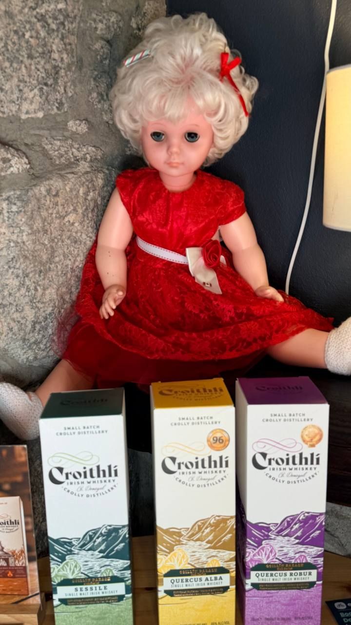 An old Crolly Doll in a red lace dress with curled white hair, seated against the granite wall beside three boxes of Croithlí whiskey.