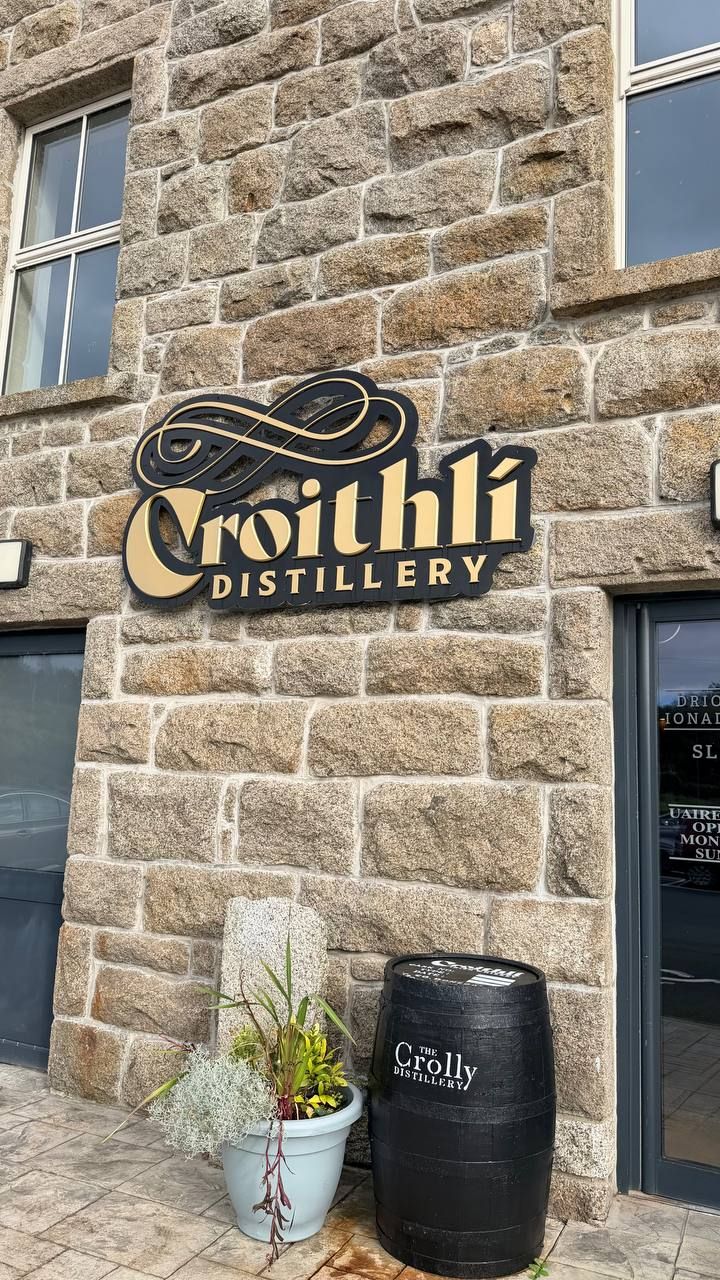 The gold and black Croithlí Distillery sign on a granite wall, with a black whiskey barrel and a pale blue flower pot below it.