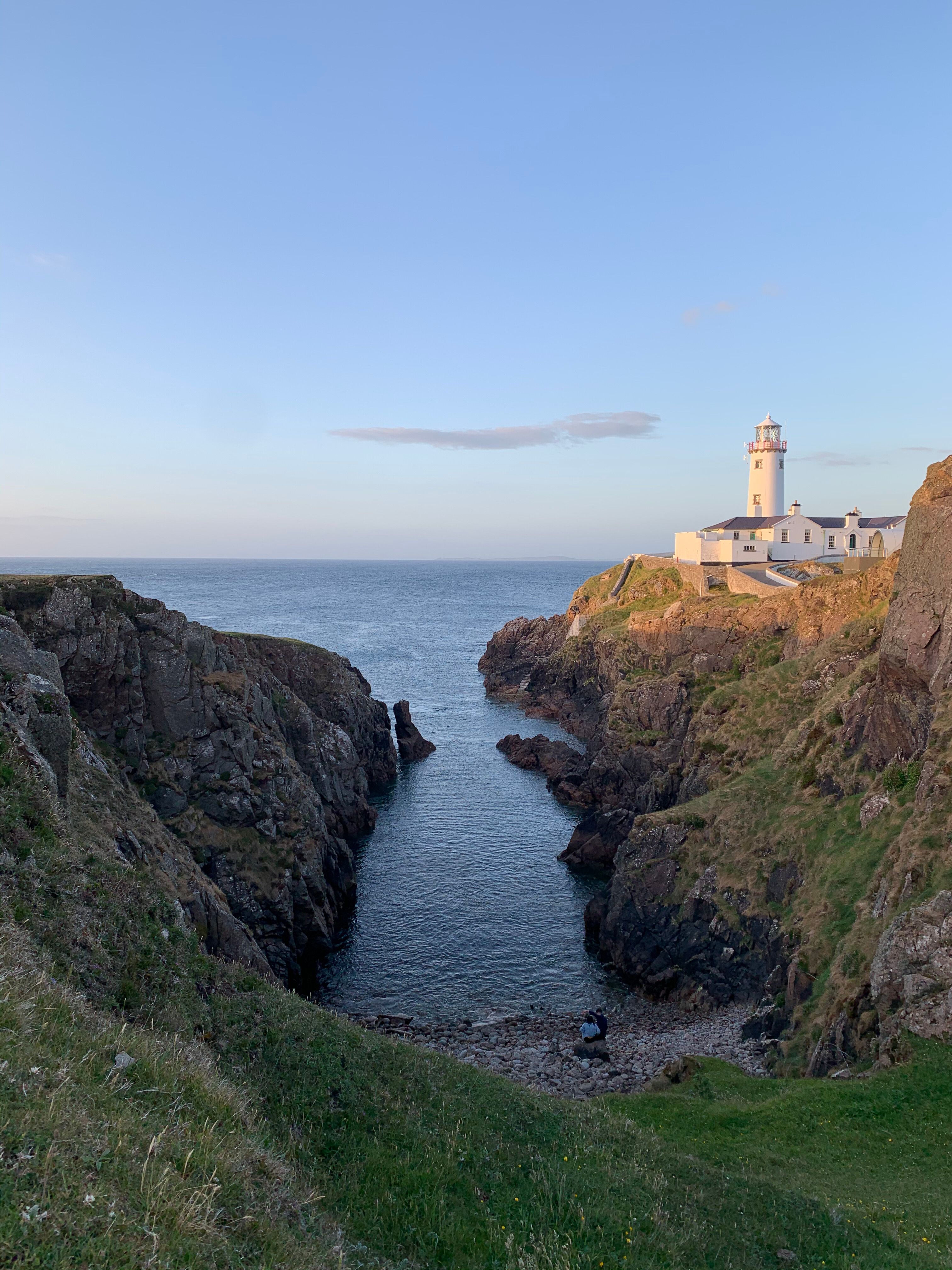 Fanad Head Lighthouse across a dramatic rocky sea inlet in golden evening light, the white tower and keeper's cottages on the headland with calm sea threading up the gorge below