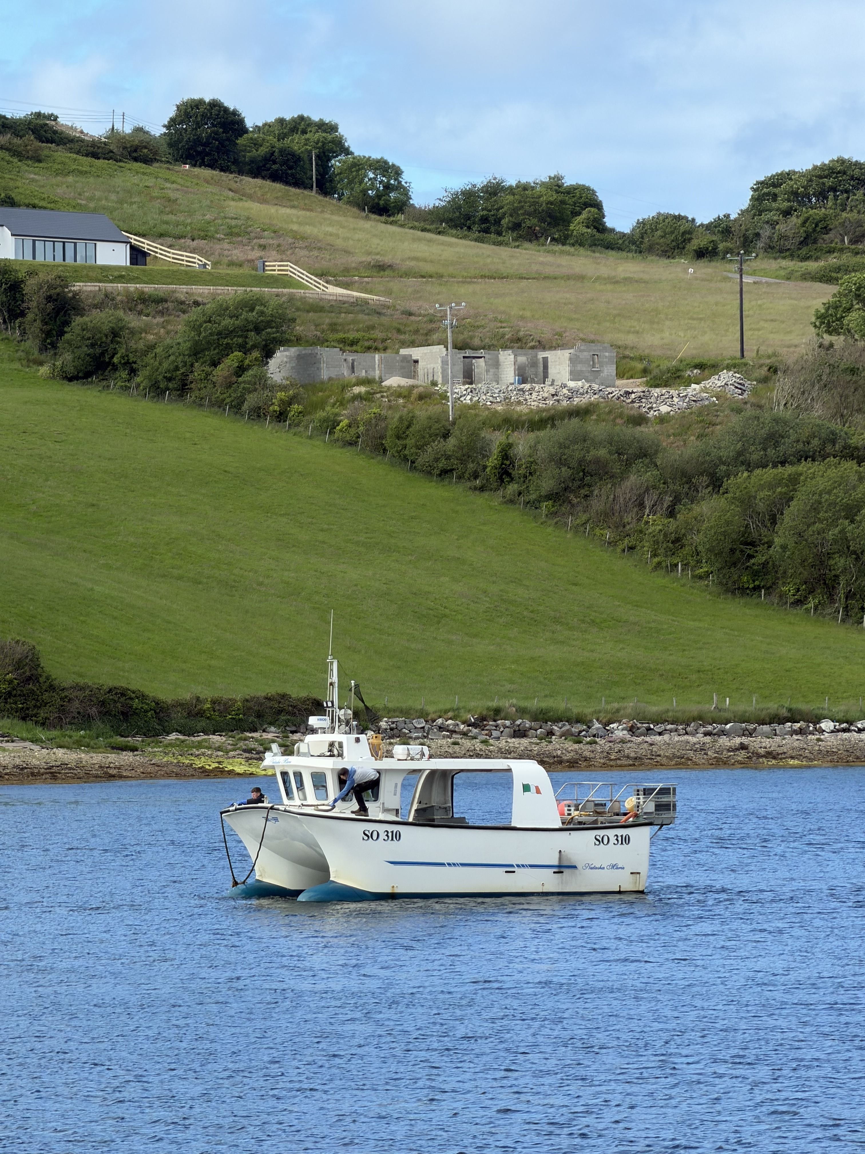 A small white inshore fishing boat, registration SO 330, moored on calm water below green fields, with a ruined stone outbuilding and a house on the hill behind.