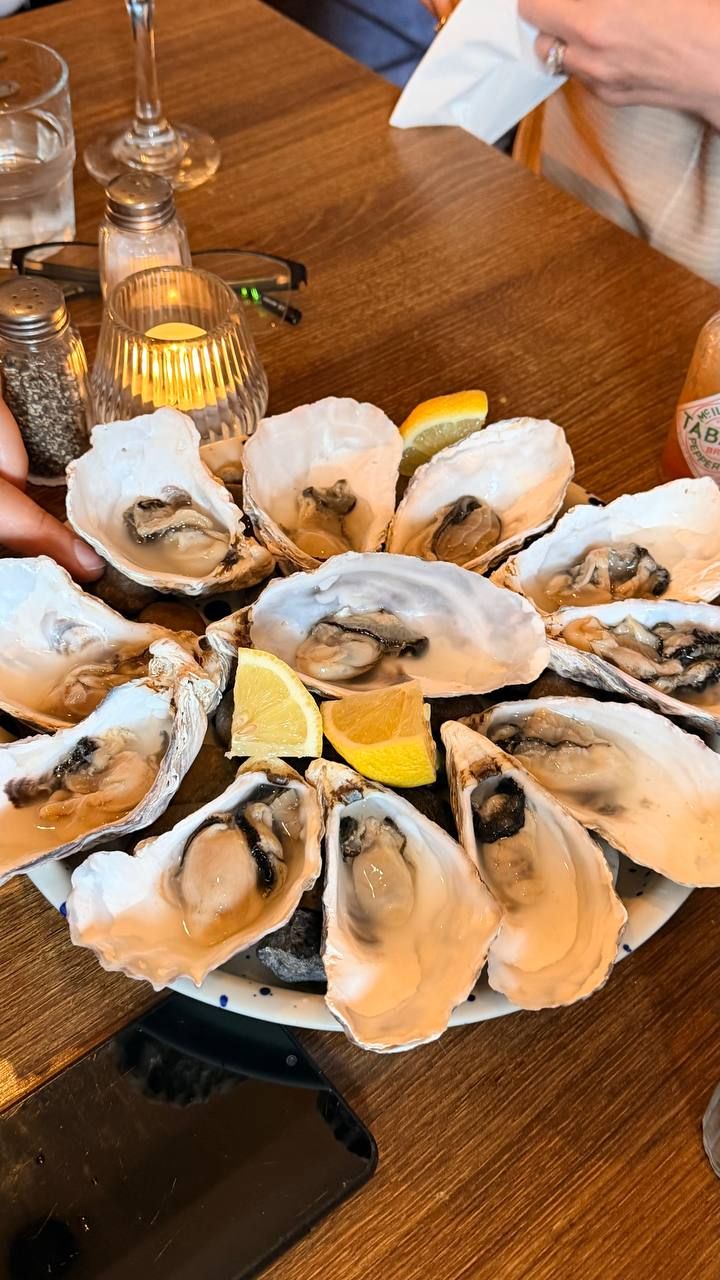 A dozen oysters on the half shell arranged in a circle on a white blue-speckled platter, lemon wedges in the middle, with a bottle of Tabasco and a lit candle behind.