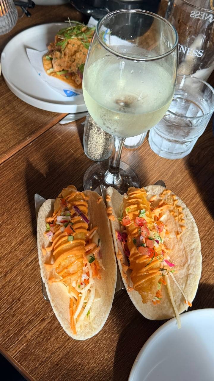 Two fish tacos in a metal stand on a wooden table at Fisk, seen from above: battered fish in soft flour tortillas with slaw, chopped tomato and red onion salsa and spiced mayonnaise, with a glass of white wine and a tumbler of water behind them.