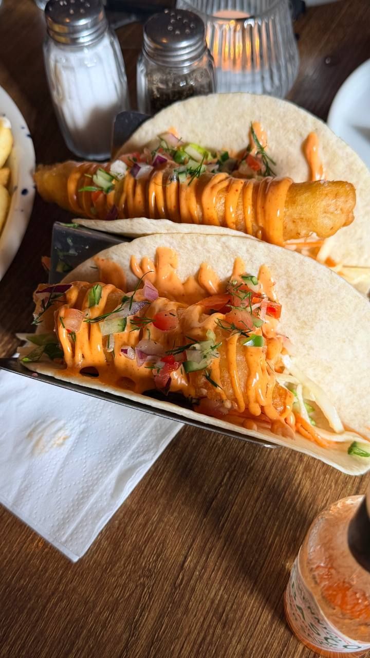 Two fish tacos close up in a metal stand: battered fish in soft flour tortillas with cabbage and carrot slaw, a chopped salsa of tomato, cucumber, red onion and dill, and a piped drizzle of pale orange spiced mayonnaise.