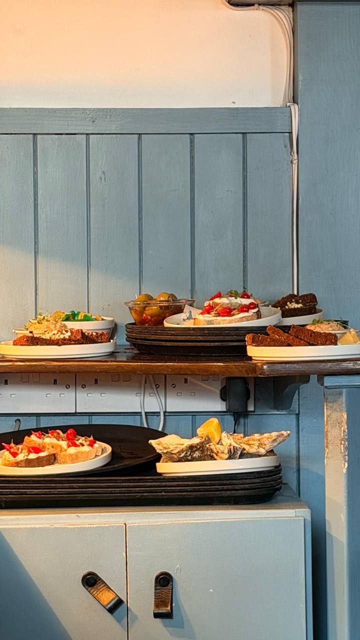 The pass inside Fisk against pale blue tongue-and-groove panelling in late evening sun: plates of oysters, sourdough toasts with whipped cheese and tomatoes, brown soda bread and a bowl of olives waiting to go out, on stacked dark serving trays.