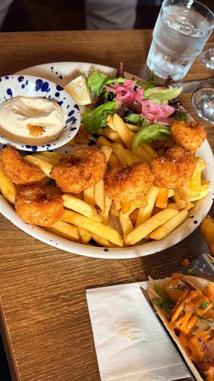 A bowl of scampi and chips: breadcrumbed prawns, golden and salted, on thick-cut chips with a leaf salad, pickled red onion, olives and a lemon wedge, with a small dish of pale aioli alongside.