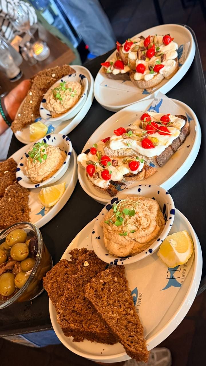 A spread of small plates on a dark table: two bowls of smoked fish pate with micro-leaves, lemon and triangles of brown soda bread, two plates of sourdough toasts topped with whipped cheese, blistered cherry tomatoes and pistachios, and a bowl of green olives.