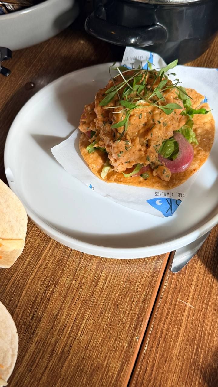 The spicy prawn cocktail tostada: prawns in a pale orange sauce flecked with herbs, over shredded lettuce on a crisp fried tostada base, with pickled red onion and a nest of micro-leaves on top, on a plate lined with a Fisk napkin.