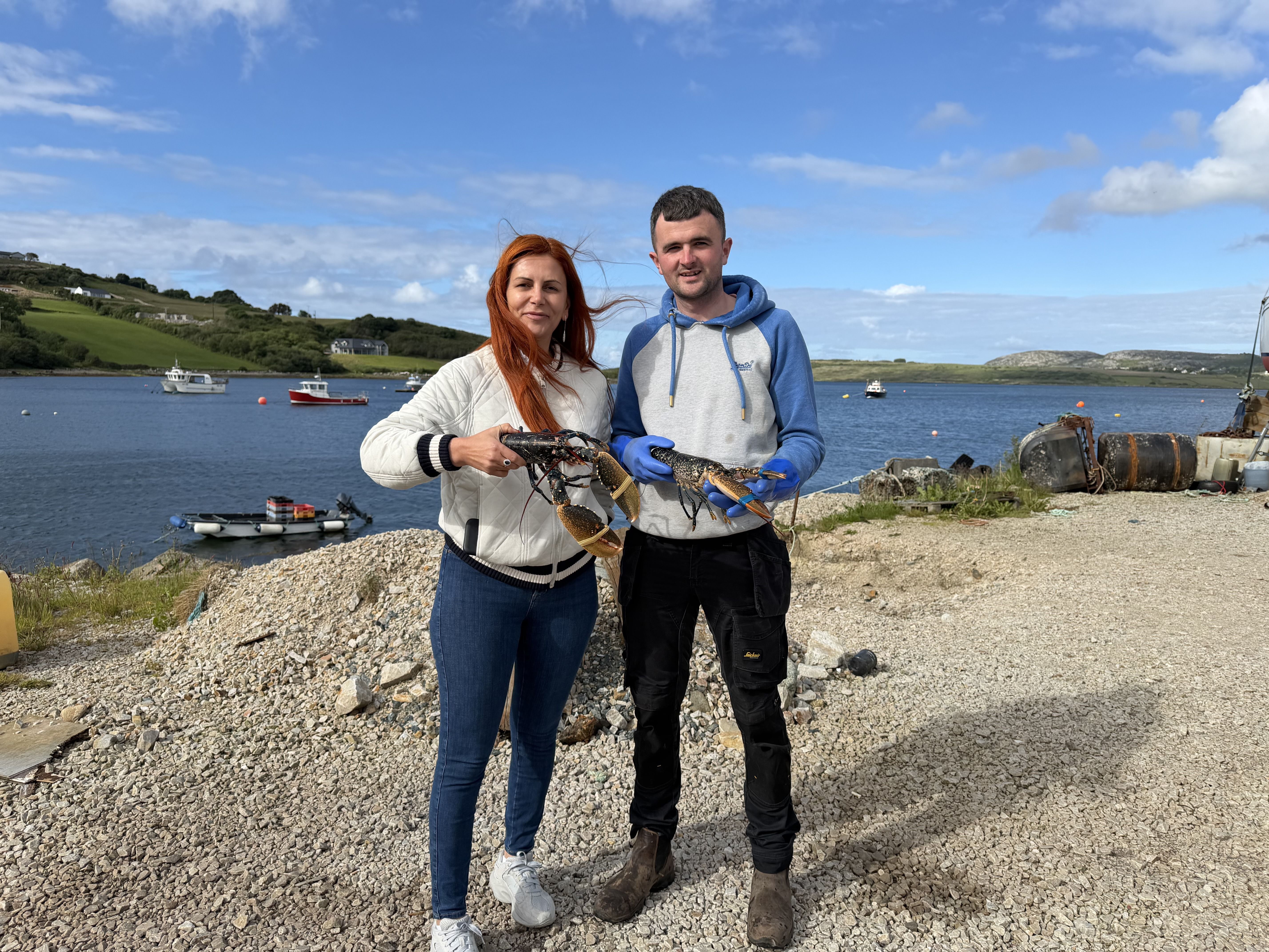 Oisin and Nihal standing on the gravel pier, each holding up a live lobster, the calm bay with moored boats and green hills behind them.