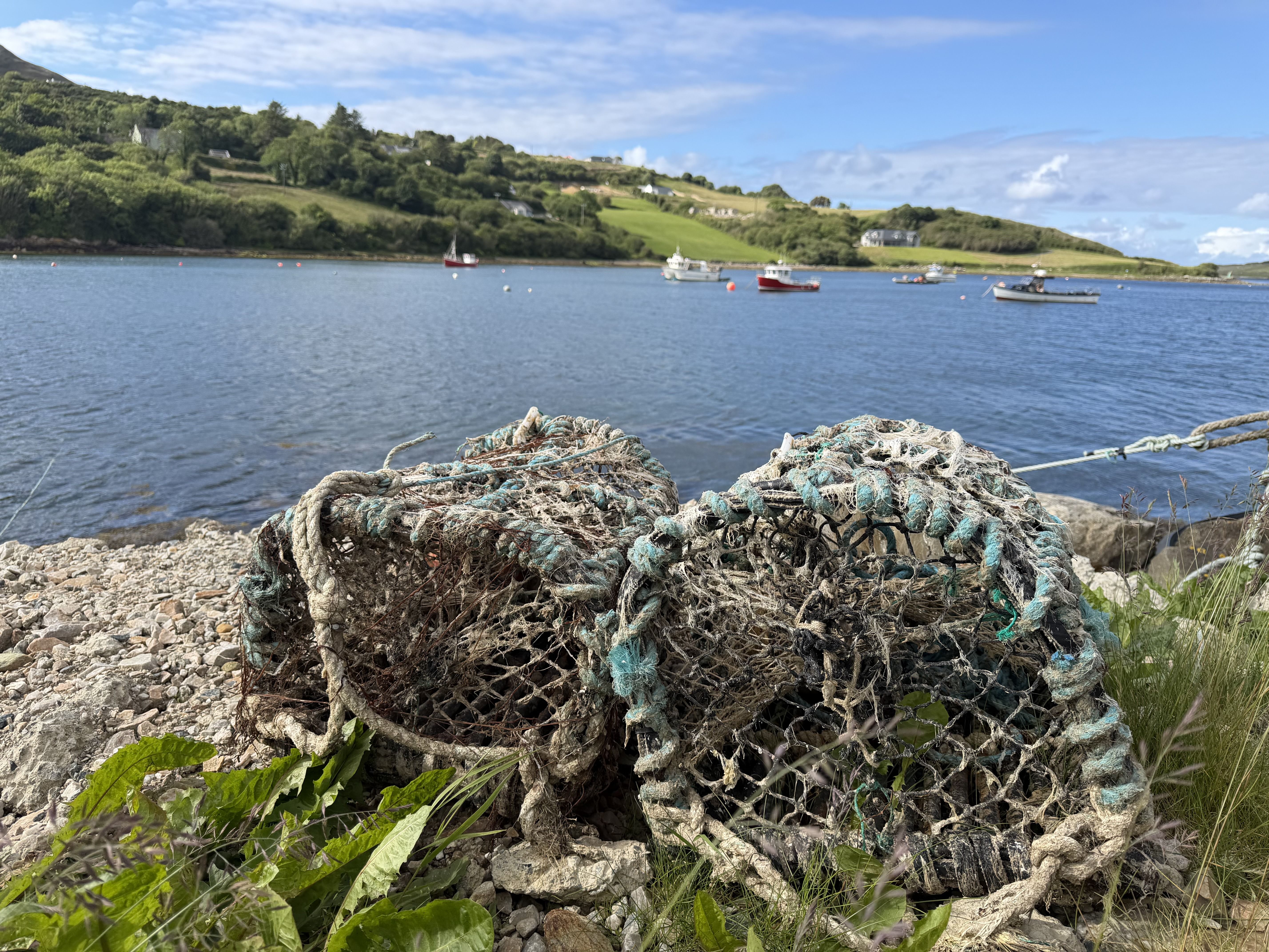 Two weathered lobster and crab creels on a rocky pier in the foreground, with moored fishing boats on a calm sea inlet beyond and green hills dotted with white houses under a blue sky.