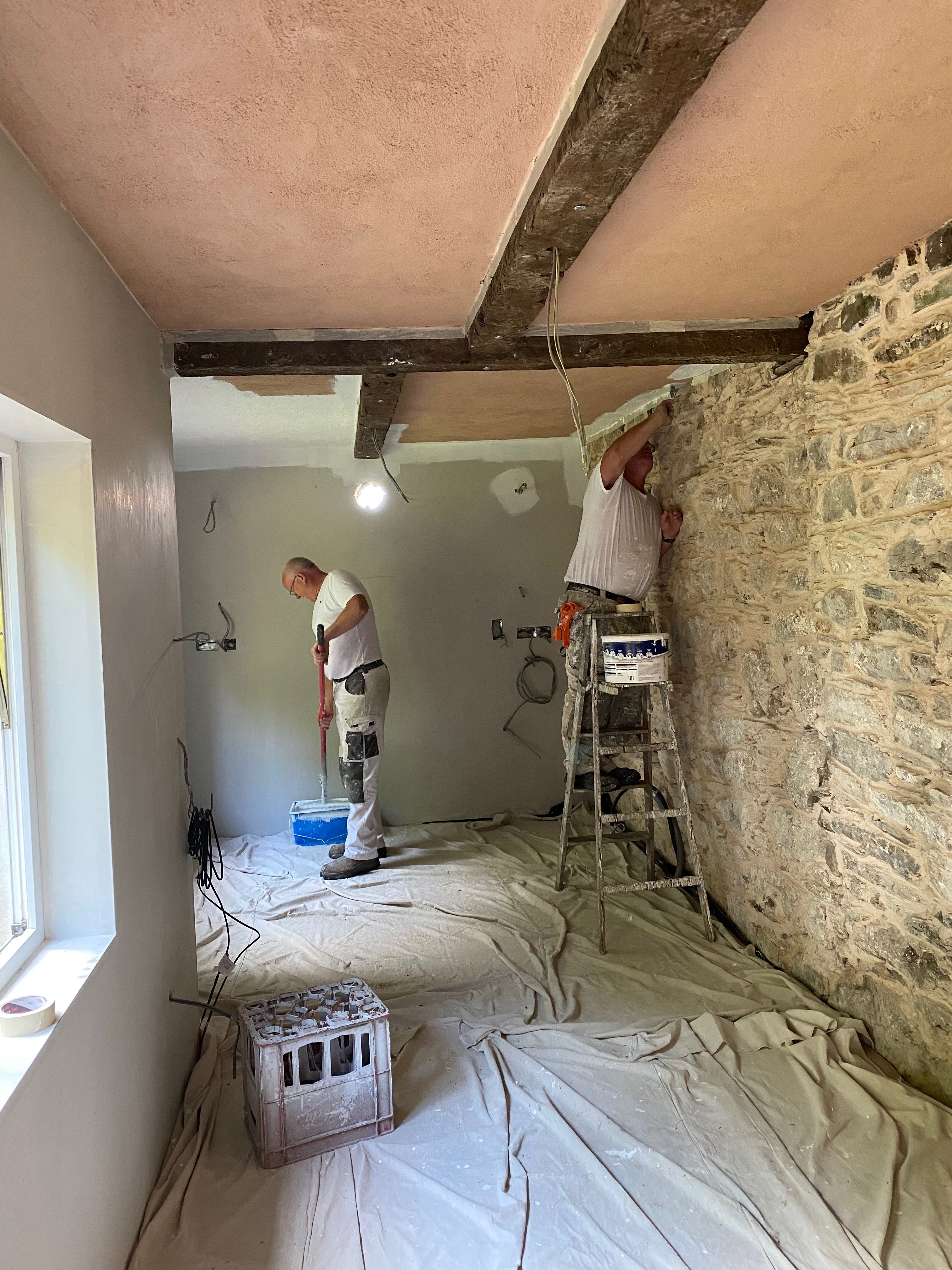 Two painters at work in the kitchen, dust sheets over the floor, the retained beam and stone wall behind.