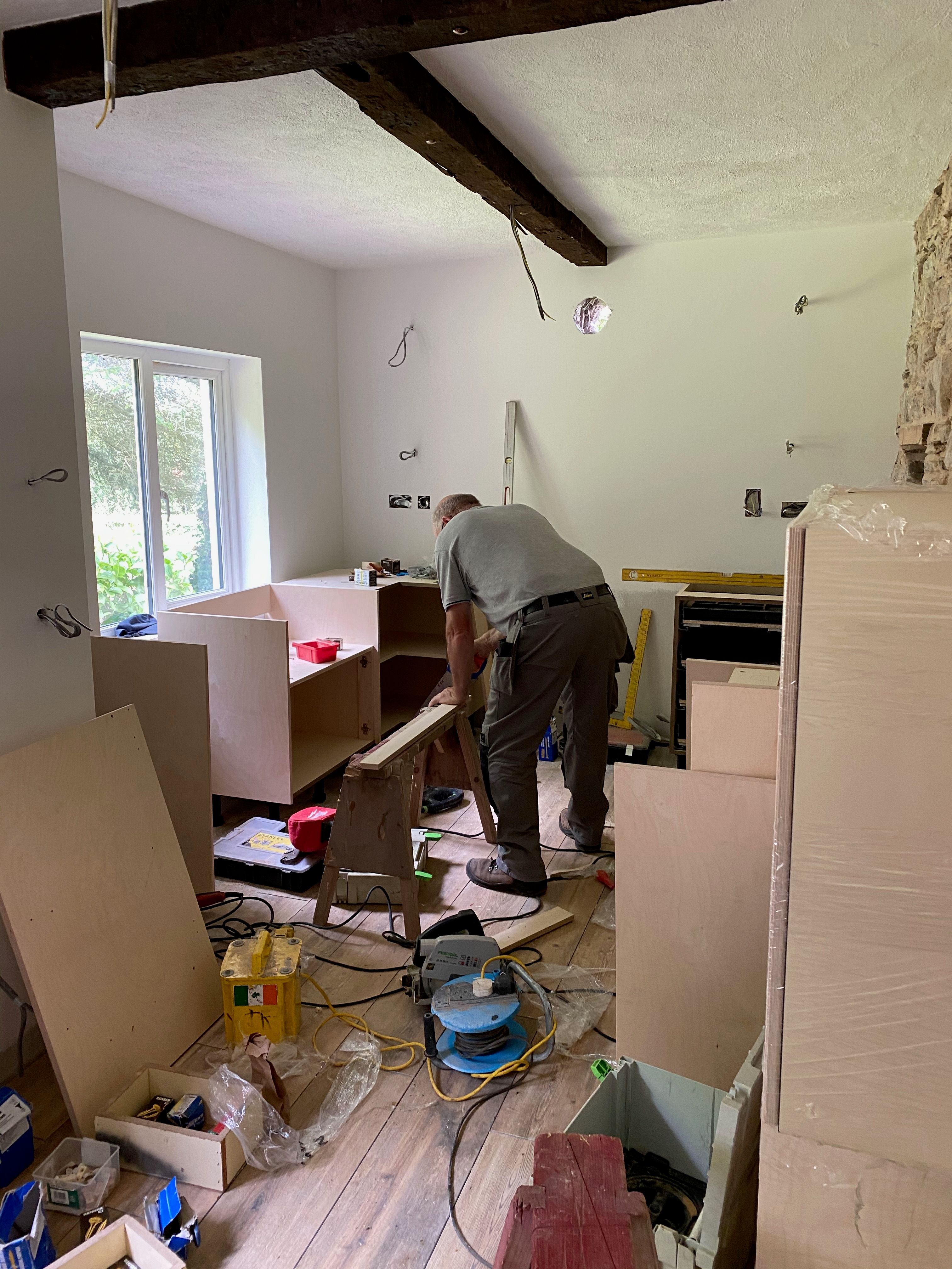 The kitchen being fitted — a carpenter cutting timber over a sawhorse amid part-built birch-ply carcasses.